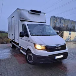Biały samochód dostawczy marki MAN z chłodnią na parkingu z kostki brukowej, w tle budynek mieszkalny i myjnia samochodowa.