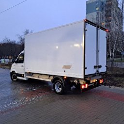 Michał Piwko - Biały samochód dostawczy z zamkniętymi drzwiami, zaparkowany na kostce brukowej, widok z tyłu, światła stopu włączone, budynek mieszkalny w tle.