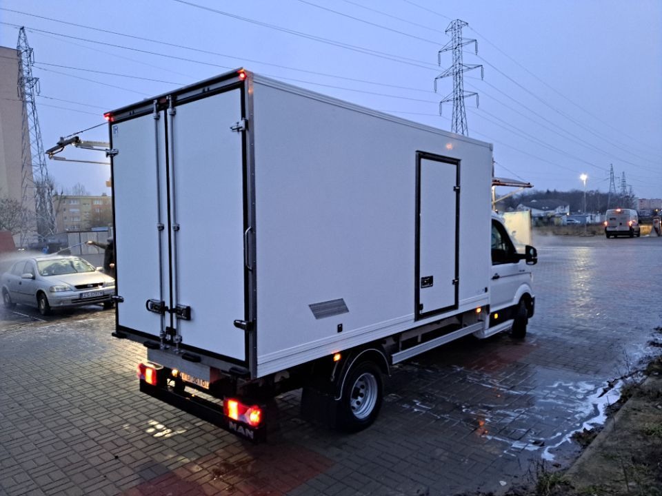 Biały samochód dostawczy MAN z zabudową kontenerową, myty na zewnątrz na mokrym placu, widok z tyłu i boku, w tle słupy wysokiego napięcia.