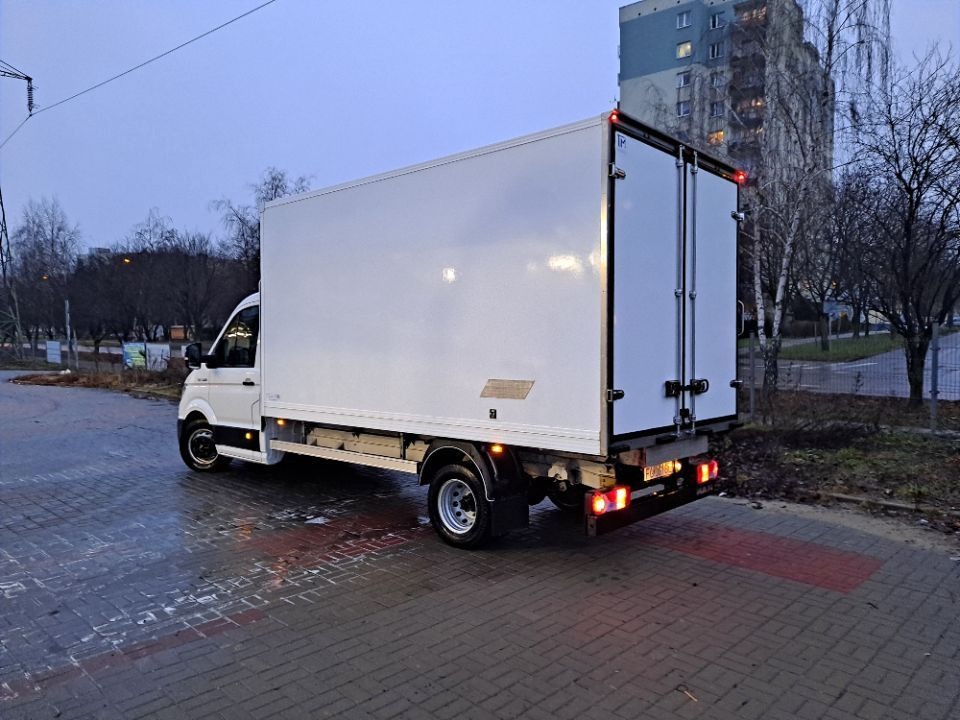 Biały samochód dostawczy z zamkniętymi drzwiami, zaparkowany na kostce brukowej, widok z tyłu, światła stopu włączone, budynek mieszkalny w tle.