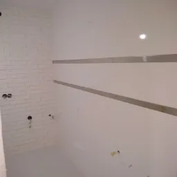 Wnętrze łazienki w trakcie remontu, widoczne białe płytki imitujące cegłę na jednej ścianie, dwie poziome listwy dekoracyjne na przeciwległej ścianie, wystające kable i rury przygotowane...