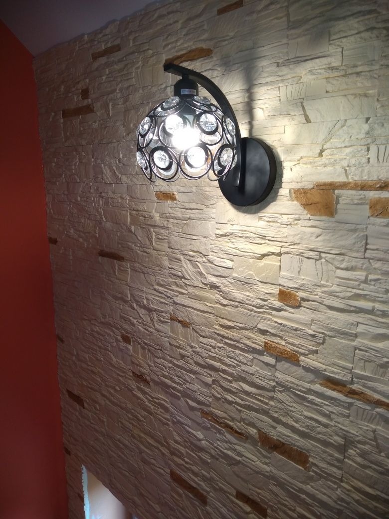 Dekoracyjna lampa ścienna z czarnym metalowym ramieniem i kloszem z kryształkami, oświetlająca ścianę wyłożoną dekoracyjnym kamieniem w odcieniach bieli i beżu.