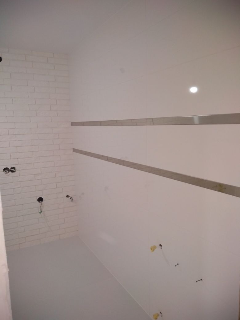 Wnętrze łazienki w trakcie remontu, widoczne białe płytki imitujące cegłę na jednej ścianie, dwie poziome listwy dekoracyjne na przeciwległej ścianie, wystające kable i rury przygotowane...