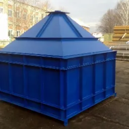 Niebieski, stalowy zbiornik z piramidalnym dachem, umieszczony na zewnątrz budynku przemysłowego, z rurami składowanymi w tle.