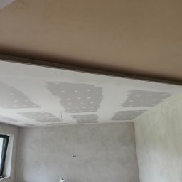 Sufit podwieszany z płyt kartonowo-gipsowych w trakcie szpachlowania łączeń, widoczne okno i fragment ściany. Prace wykończeniowe w jasnym wnętrzu.