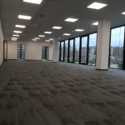 Pusta przestrzeń biurowa z szarą wykładziną dywanową, dużymi oknami z widokiem na budynki i równomiernym oświetleniem sufitowym.