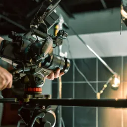 Profesjonalna kamera filmowa na statywie, obsługiwana przez operatora w studiu z oświetleniem. Widoczny duży obiektyw i elementy sterujące. Tło stanowią płytki ceramiczne.