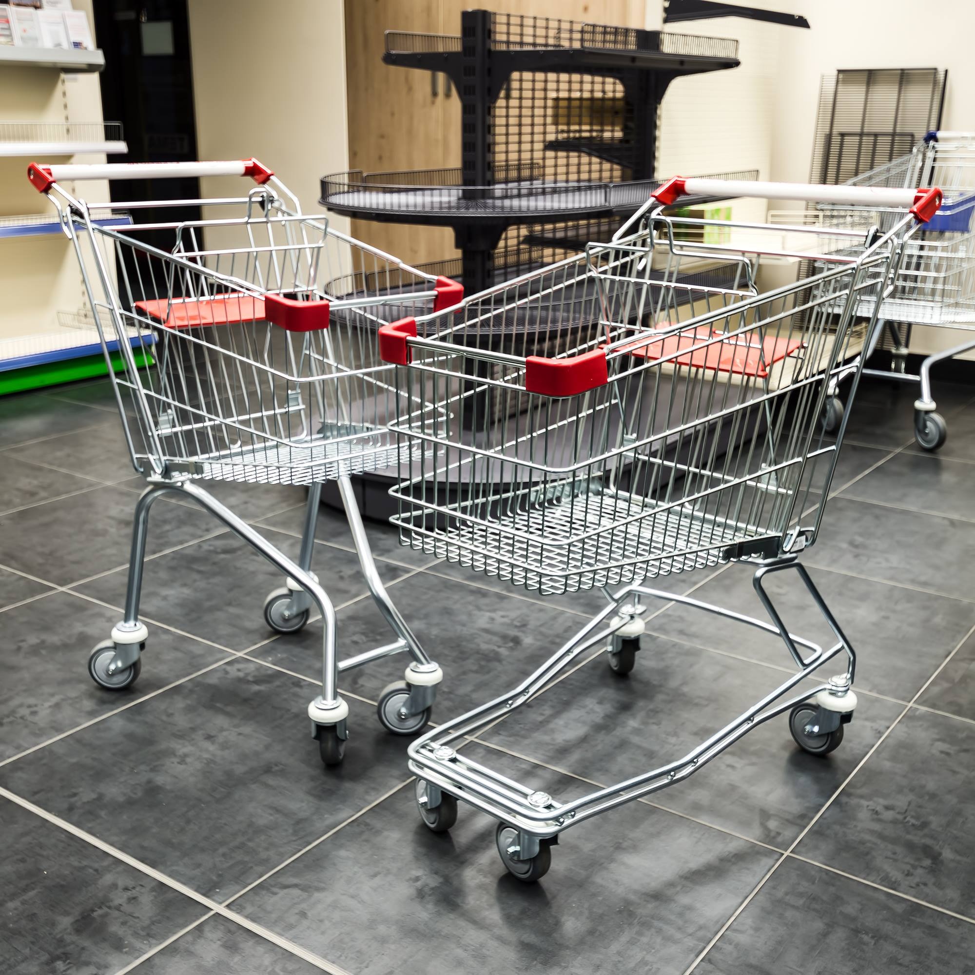 Dwa chromowane wózki sklepowe z czerwonymi uchwytami i siedziskami, stojące na ciemnej podłodze z płytek, z widocznymi regałami sklepowymi w tle.