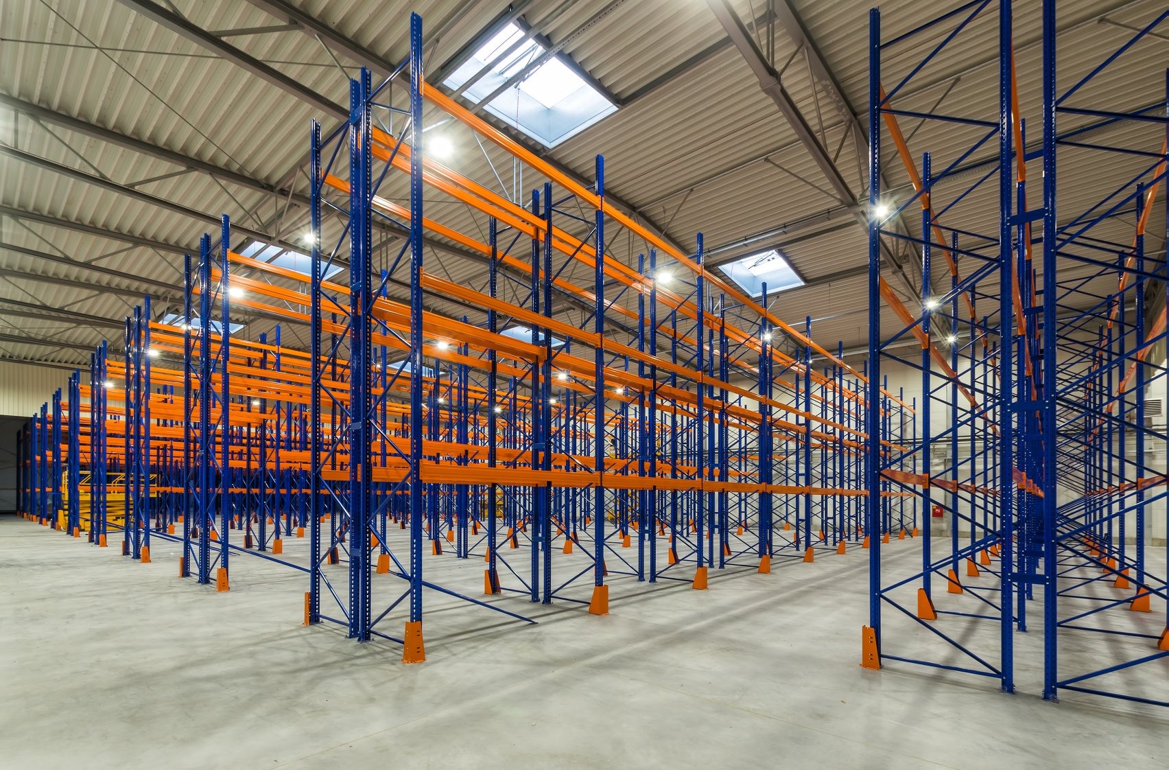 Puste, niebiesko-pomarańczowe metalowe regały magazynowe w hali z betonową podłogą i oświetleniem sufitowym.