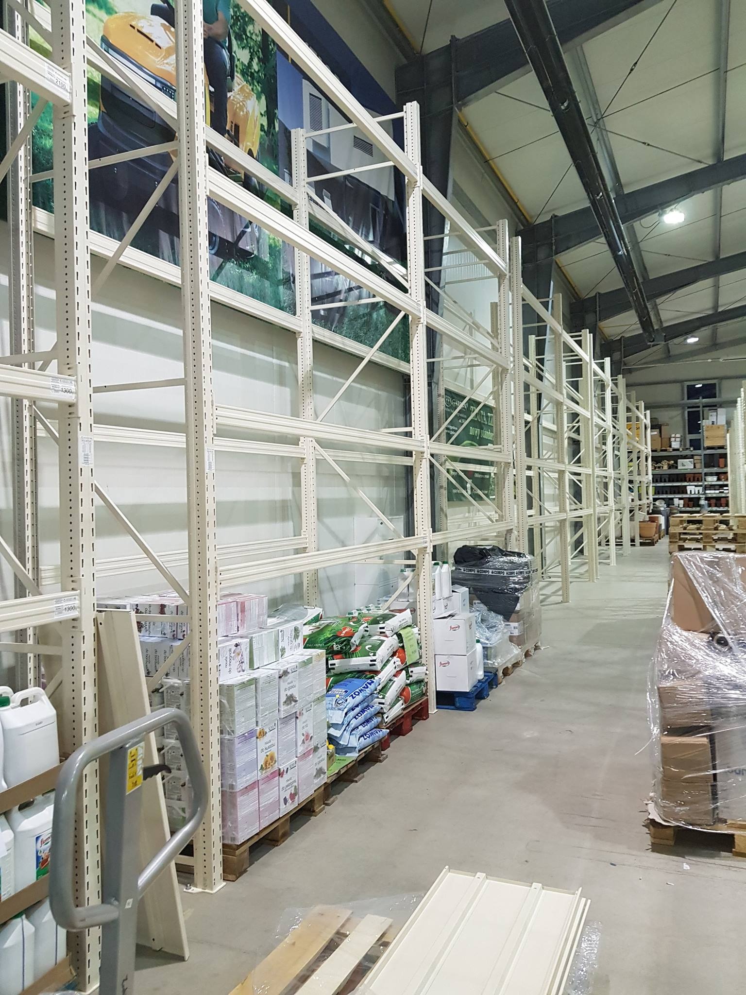 Długi rząd pustych, metalowych regałów w magazynie, częściowo wypełnionych towarem, z widoczną grafiką na ścianie w tle.