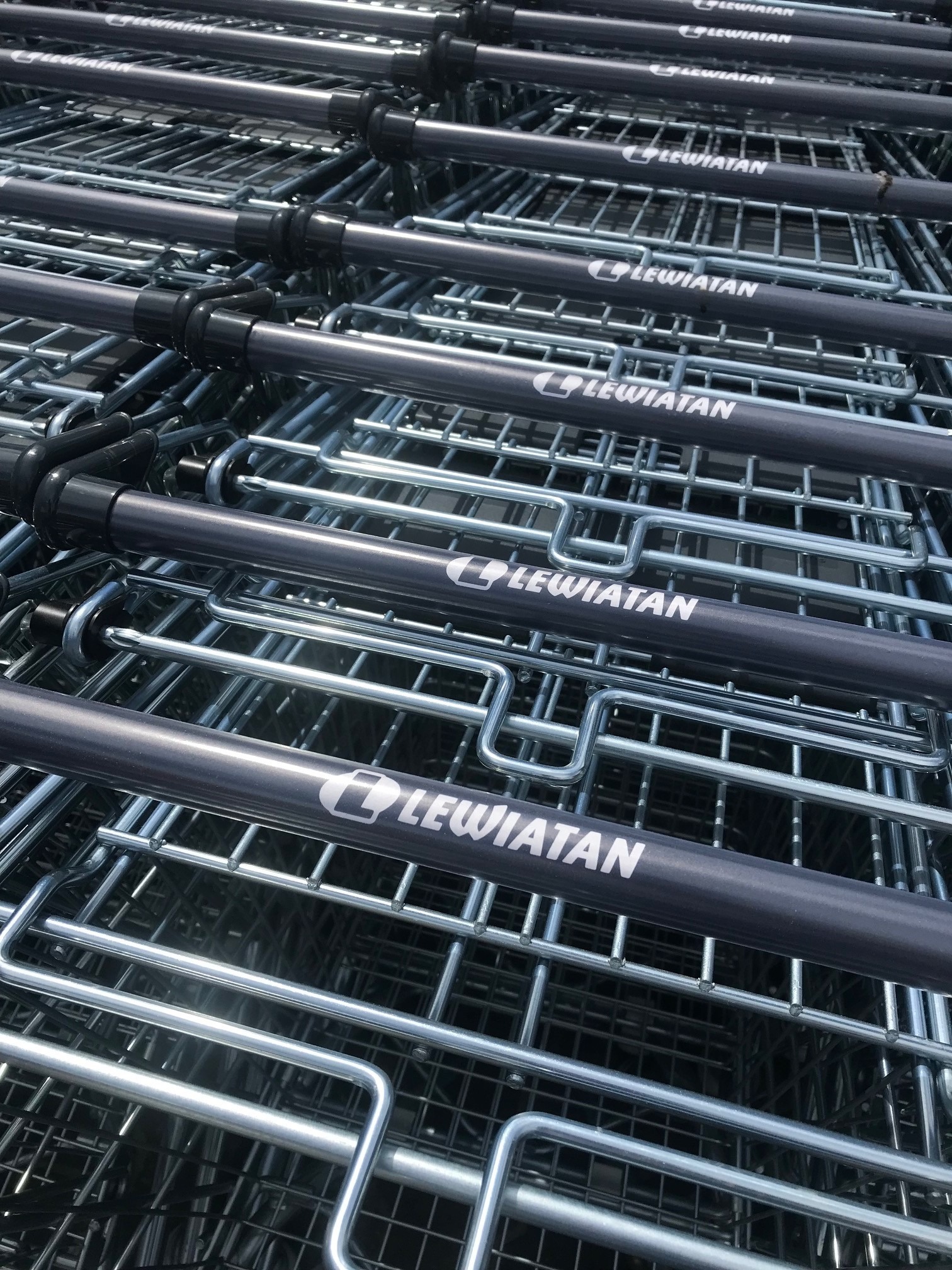 Stos metalowych wózków sklepowych z szarymi uchwytami i białym logo 'Lewiatan' na każdym uchwycie, ułożonych jeden w drugim.