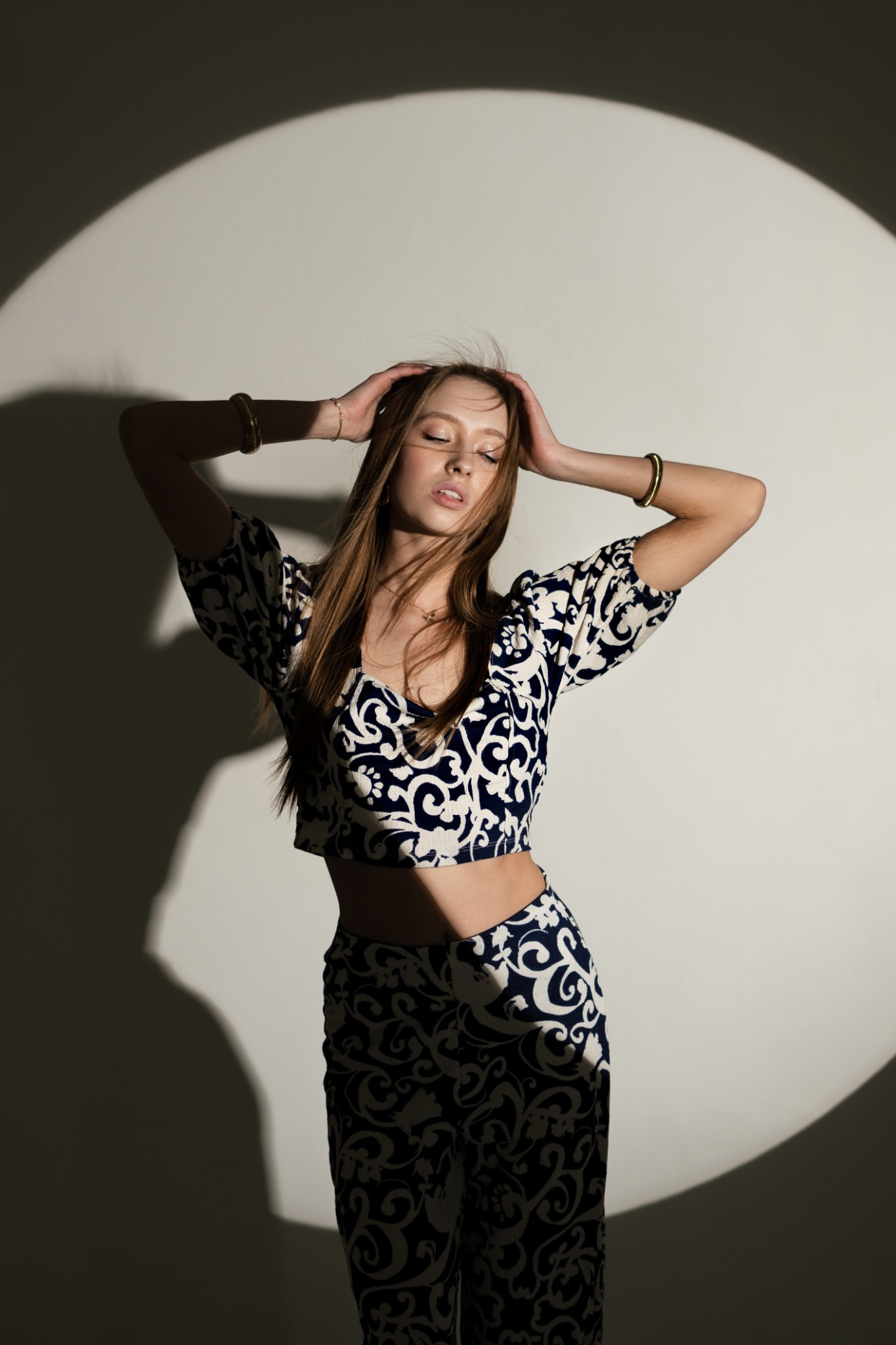 Młoda kobieta w dwuczęściowym stroju z wzorem paisley, pozuje z zamkniętymi oczami na tle oświetlonej ściany. Na rękach ma złote bransoletki.