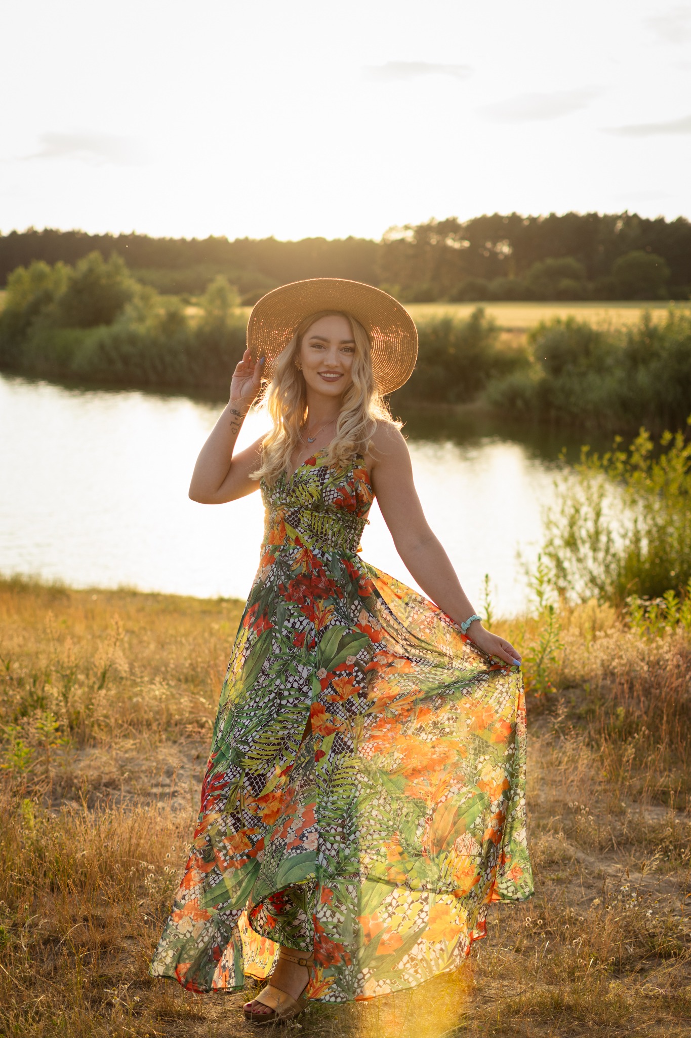Uśmiechnięta blondynka w kwiecistej sukience i kapeluszu na tle jeziora w promieniach zachodzącego słońca, trzymająca brzeg sukienki. Romantyczna sceneria.