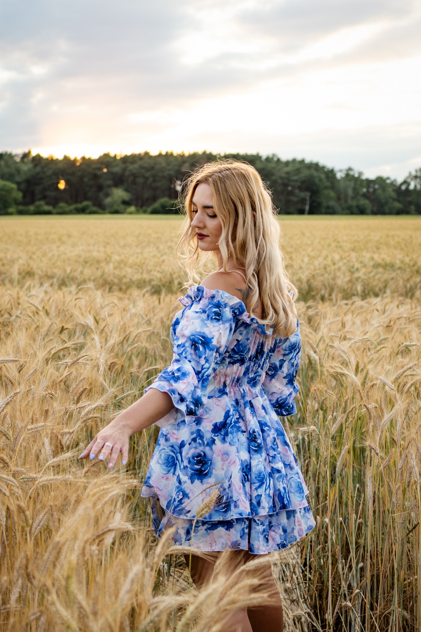 Kobieta w kwiecistej sukience, stojąca na polu pszenicy, dotyka kłosów. W tle las i zachodzące słońce, tworzące ciepłe, naturalne światło. Romantyczna sesja na łonie natury.