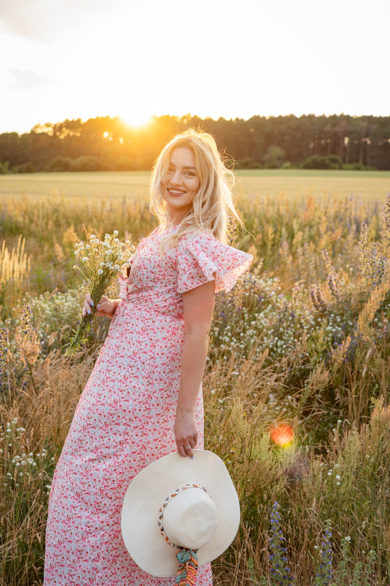 Uśmiechnięta blondynka w kwiecistej sukience na tle łąki o zachodzie słońca trzyma kapelusz i bukiecik polnych kwiatów. Ciepłe światło, naturalne otoczenie.