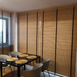 Jasne żaluzje bambusowe z czarnymi taśmami na oknach w jadalni, z widocznym stołem i krzesłami. Dają ciepłe światło do wnętrza. Sterowanie elektryczne.