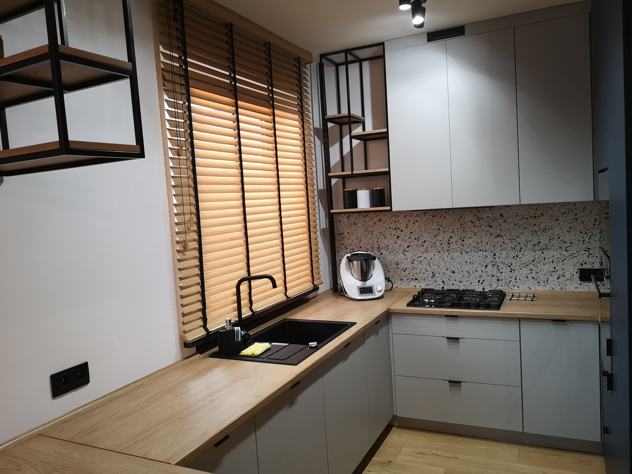 Bambusowe żaluzje z czarnymi taśmami w nowoczesnej kuchni. Żaluzje zamontowane w oknie nad zlewem, z widocznym blatem i elementami wystroju.