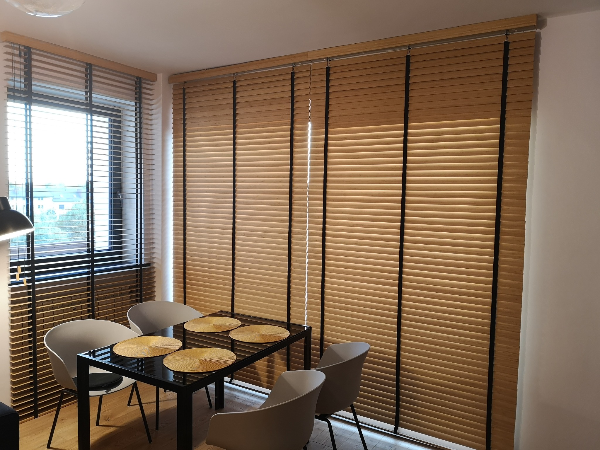 Jasne żaluzje bambusowe z czarnymi taśmami na oknach w jadalni, z widocznym stołem i krzesłami. Dają ciepłe światło do wnętrza. Sterowanie elektryczne.
