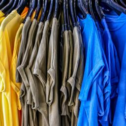 Mc Studio Reklamowe - Rząd żółtych, szarych i niebieskich koszulek z krótkim rękawem wiszących na czarnych wieszakach w sklepie odzieżowym.