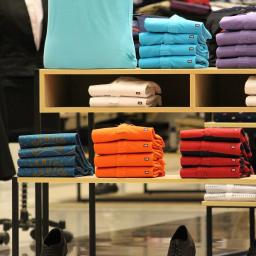 Mc Studio Reklamowe - Uporządkowane stosy różnokolorowych koszul męskich i damskich na drewnianych półkach w sklepie odzieżowym, z garniturami w tle i butami na podłodze.