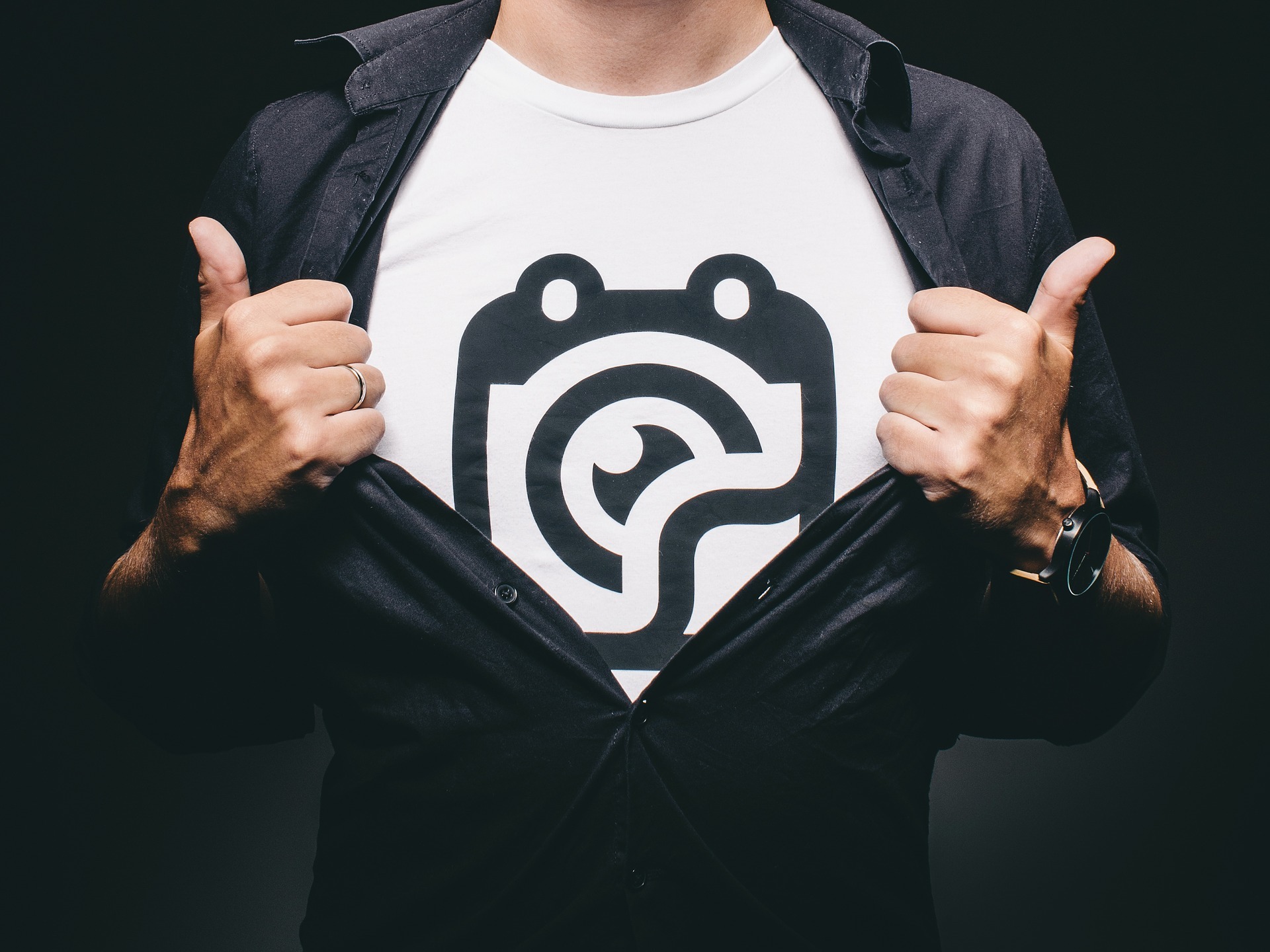 Mężczyzna rozpinający czarną koszulę, odsłaniając biały T-shirt z dużym, czarnym logo. Widoczny pierścionek na palcu i zegarek na ręce. Zdjęcie wykonane w Sulechowie.