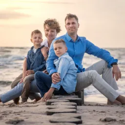 Rodzina z dwójką dzieci siedzi na drewnianych palach na plaży, morze w tle, ciepłe światło zachodzącego słońca.