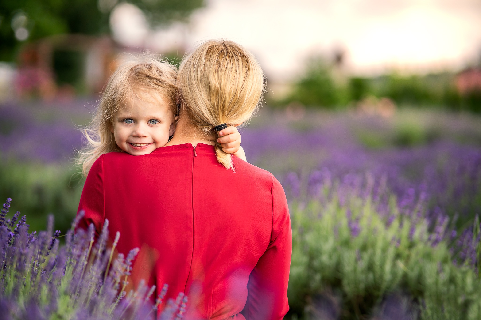 Uśmiechnięta blond dziewczynka wtulona w szyję kobiety w czerwonej sukience na tle pola lawendy.