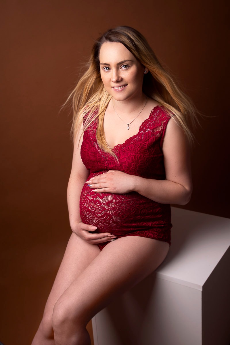 Portret kobiety w ciąży, ubranej w koronkową bieliznę w odcieniach czerwieni, siedzącej na białym postumencie na tle brązowego tła, delikatnie uśmiechniętej.