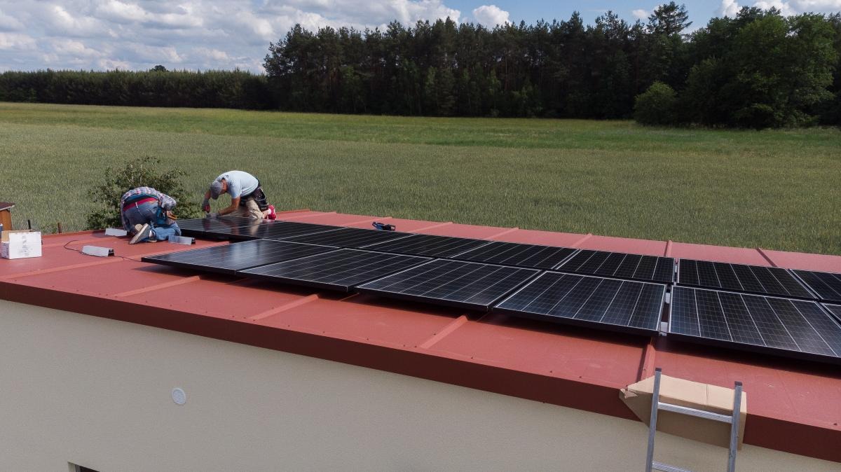 Montaż paneli słonecznych na czerwonym dachu w Warszawie, widoczne dwie osoby pracujące oraz zielone pole w tle.