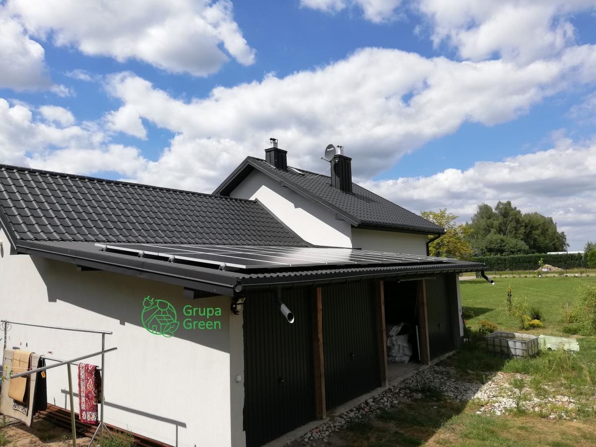 Panele słoneczne zamontowane na dachu garażu przylegającego do domu jednorodzinnego, widoczne logo firmy 'Grupa Green' na ścianie, suszarka z dywanami z boku garażu.