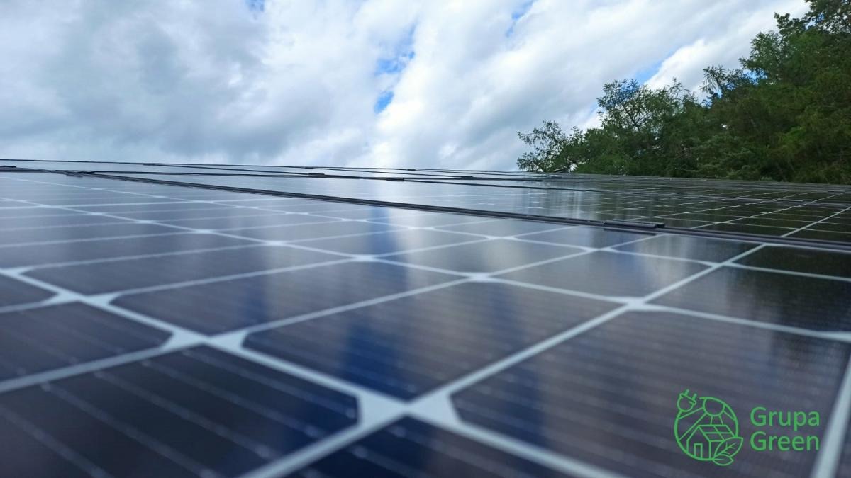 Panel słoneczny na dachu budynku, odbijający pochmurne niebo, z widocznym logo firmy Grupa Green w prawym dolnym rogu.
