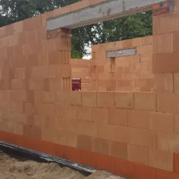 Wznoszone ściany z pustaków ceramicznych Porotherm z widocznymi nadprożami okiennymi i izolacją przeciwwilgociową fundamentów na etapie budowy domu, w tle drzewa.