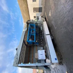 Wnętrze skrzyni ładunkowej busa z niebieską maszyną rolniczą i paletą kartonów z napojami, widok z tyłu przy otwartych drzwiach, tło: budynek i niebo.