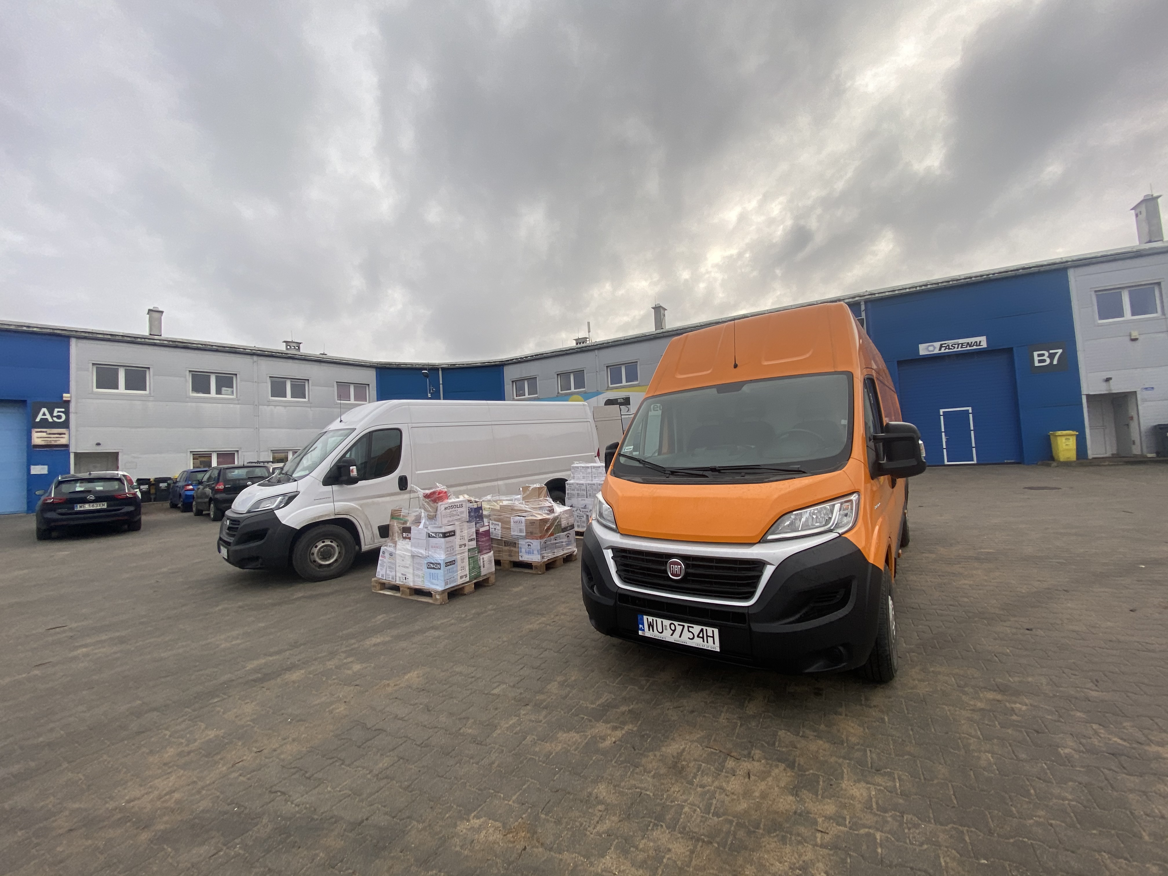 Dwa busy: biały i pomarańczowy, zaparkowane na placu przed budynkiem magazynowym, palety z zapakowanymi towarami między nimi, pochmurne niebo.