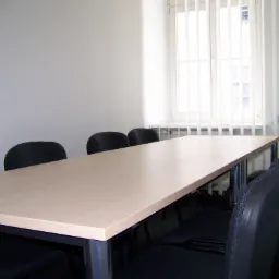 Sala konferencyjna z długim stołem z jasnego drewna i rzędem czarnych krzeseł na metalowych nogach, jasne tło z oknem z białymi żaluzjami.