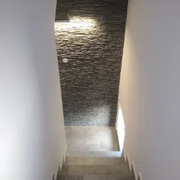 Wykończenie ściany schodów kamieniem dekoracyjnym w odcieniach szarości, z podświetleniem LED i szarymi płytkami na schodach.