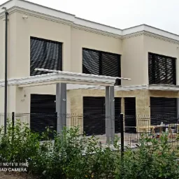 Elewacja dwukondygnacyjnego domu z beżową fasadą, czarnymi roletami zewnętrznymi i dekoracyjnymi elementami architektonicznymi, widziana zza metalowego ogrodzenia i krzewów.