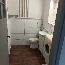 Biała łazienka z brązową podłogą imitującą drewno, widoczna toaleta, umywalka z szafką, pralka, grzejnik żeberkowy i częściowo otwarte drzwi.