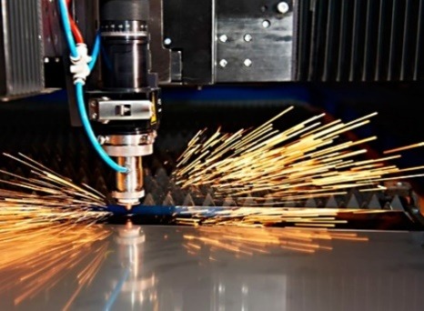 Precyzyjne cięcie metalu laserem przemysłowym, iskry rozbryzgują się wokół, widoczne elementy maszyny i obrabiany arkusz blachy.