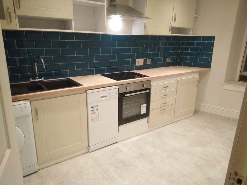 Nowoczesna kuchnia z ciemnoturkusową glazurą ścienną ułożoną w cegiełkę, jasnymi szafkami i drewnianym blatem, wyposażona w czarny zlew, płytę indukcyjną i sprzęt AGD.