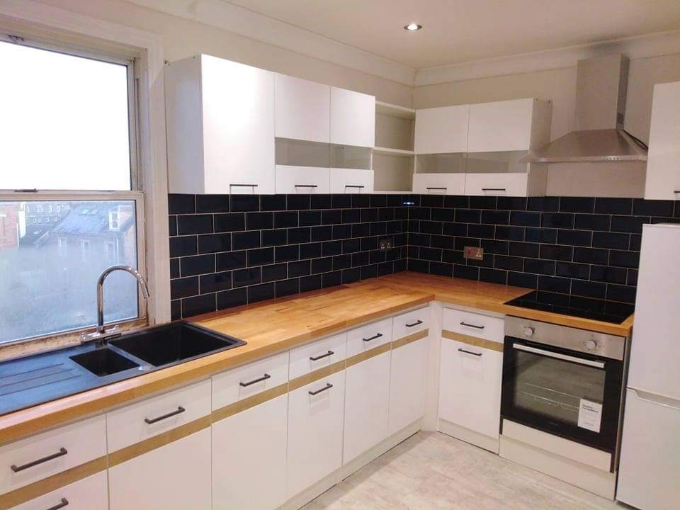 Nowoczesna kuchnia z białymi szafkami, drewnianym blatem i czarną glazurą na ścianie, widok na zewnątrz przez okno.