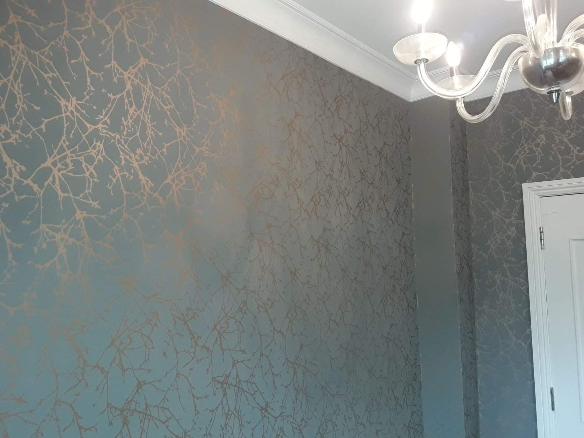 Ściana w kolorze gołębim z delikatnym, metalicznym wzorem gałązek, wykończona białym, ozdobnym gzymsem sufitowym i widocznym fragmentem kryształowego żyrandola.