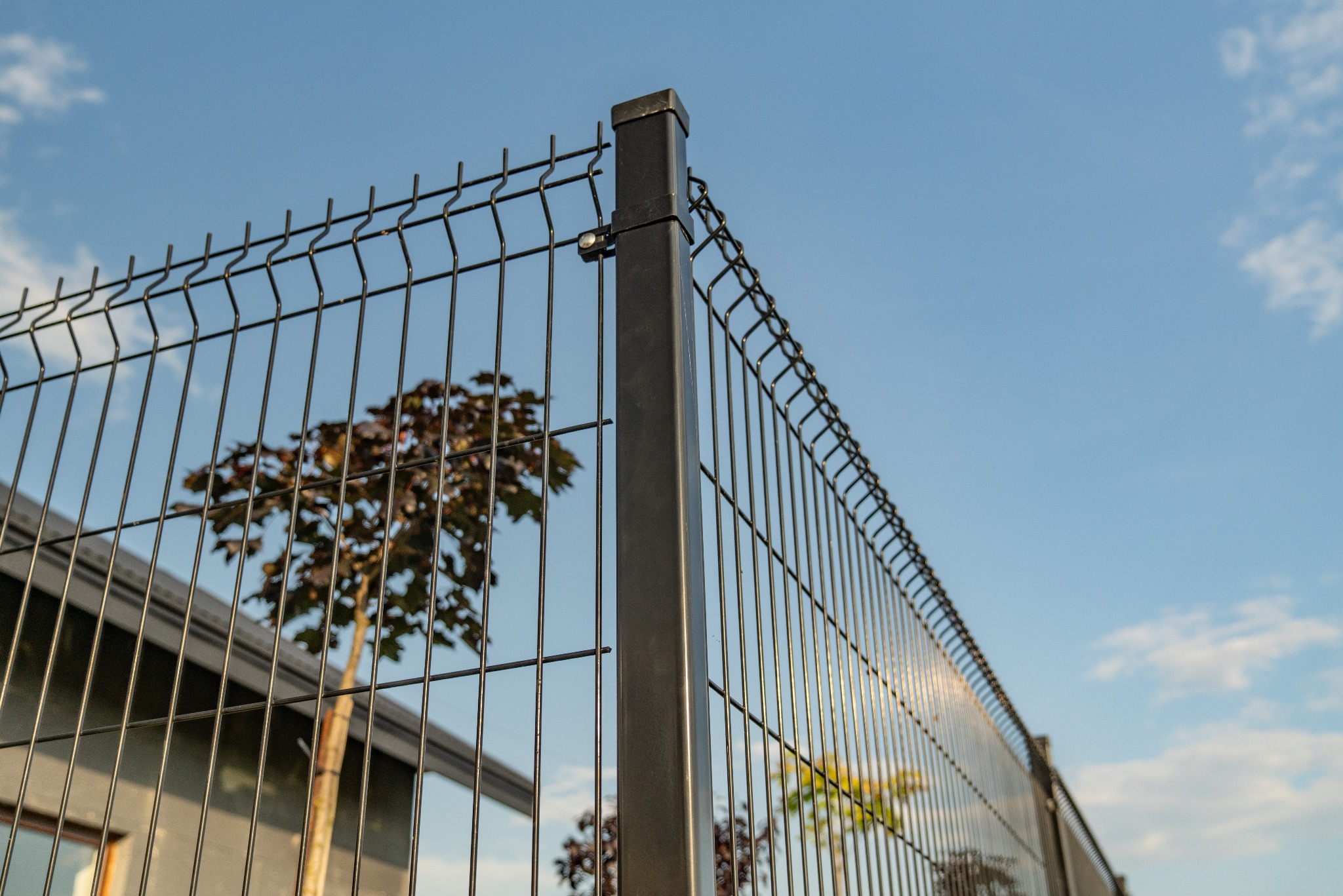 Szary panel ogrodzeniowy z widocznym słupkiem mocującym, na tle błękitnego nieba i młodego drzewka o bordowych liściach, przylegający do nowoczesnego budynku.