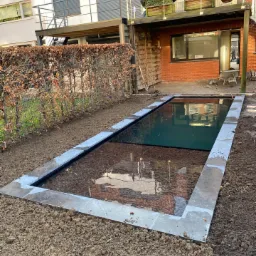 Prostokątny zbiornik wodny z kamienną obudową w trakcie budowy, z widocznym dnem wyłożonym kamieniami i częściowo wypełniony wodą, otoczony ziemią i żywopłotem, na tle nowoczesnego domu z balkonem.