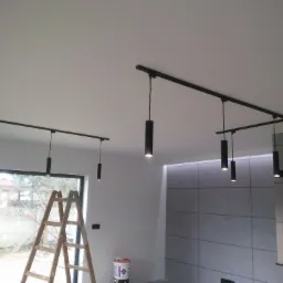 Nowoczesne czarne lampy tuby na szynoprzewodzie zamontowane na suficie, widoczna drabina i wiaderko z farbą sugerujące etap wykończeniowy.