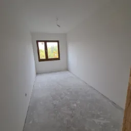 Pusty pokój po nałożeniu gładzi, z widocznym oknem i surowym betonem na podłodze, przygotowany do dalszych prac wykończeniowych.