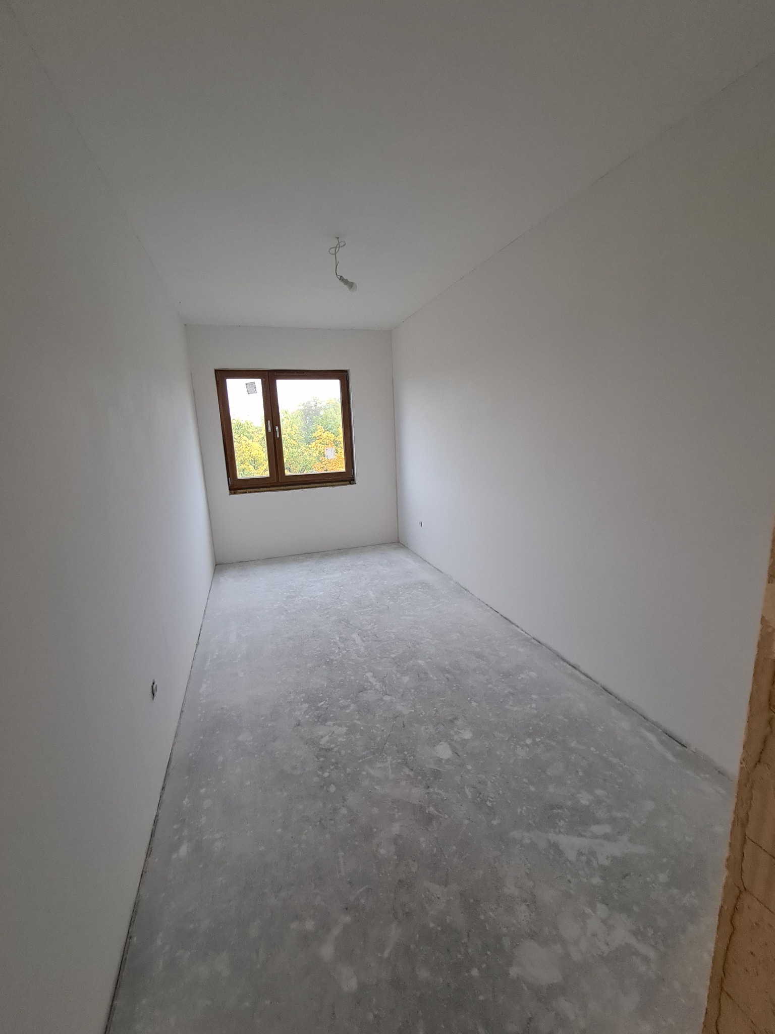 Pusty pokój po nałożeniu gładzi, z widocznym oknem i surowym betonem na podłodze, przygotowany do dalszych prac wykończeniowych.