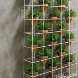 Metalowa konstrukcja w formie kraty, pomalowana na biało, służąca jako pionowy kwietnik, z doniczkami z zielonymi roślinami i kamieniami na dole, umieszczona na tle szarej ściany.