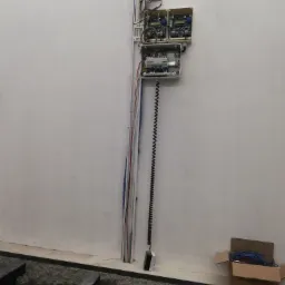 Zamontowana na białej ścianie skrzynka z elektroniką, widoczne kable i przewody, na dole czarny kabel spiralny i karton z niebieskimi kablami.