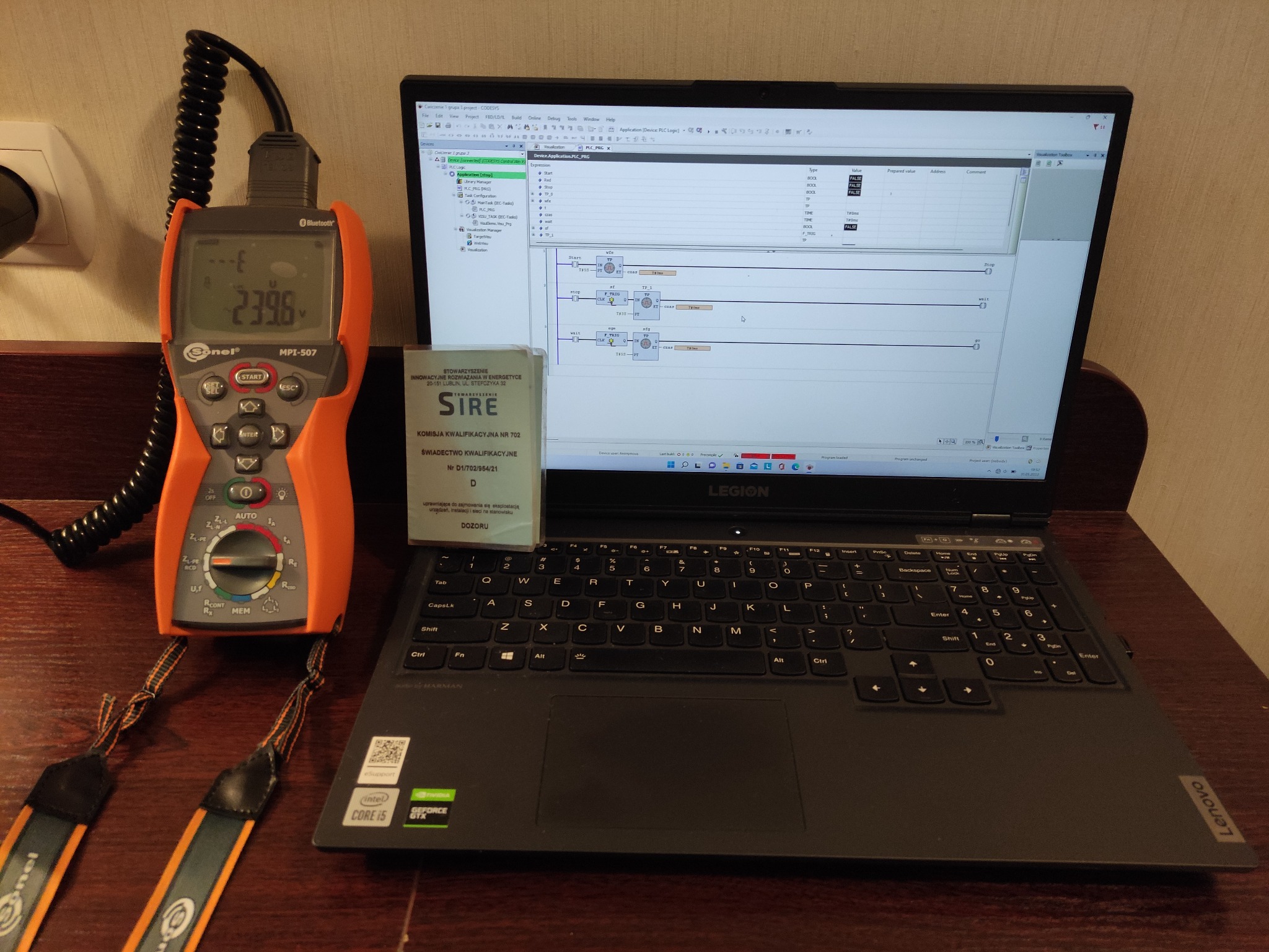 Pomarańczowy miernik Sonel MPI-507 z wyświetlanym napięciem 239.6V, leżący obok laptopa Lenovo Legion z uruchomionym programem do sterowania PLC i dokumentem kwalifikacyjnym SIRE na klawiaturze.